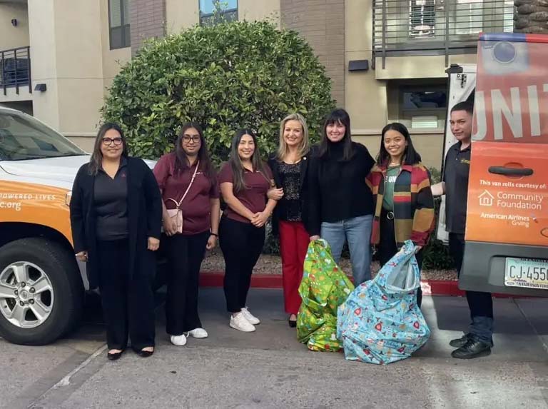 Group of volunteers standing outdoors beside a United Way vehicle, holding large donation bags in front of an apartment building